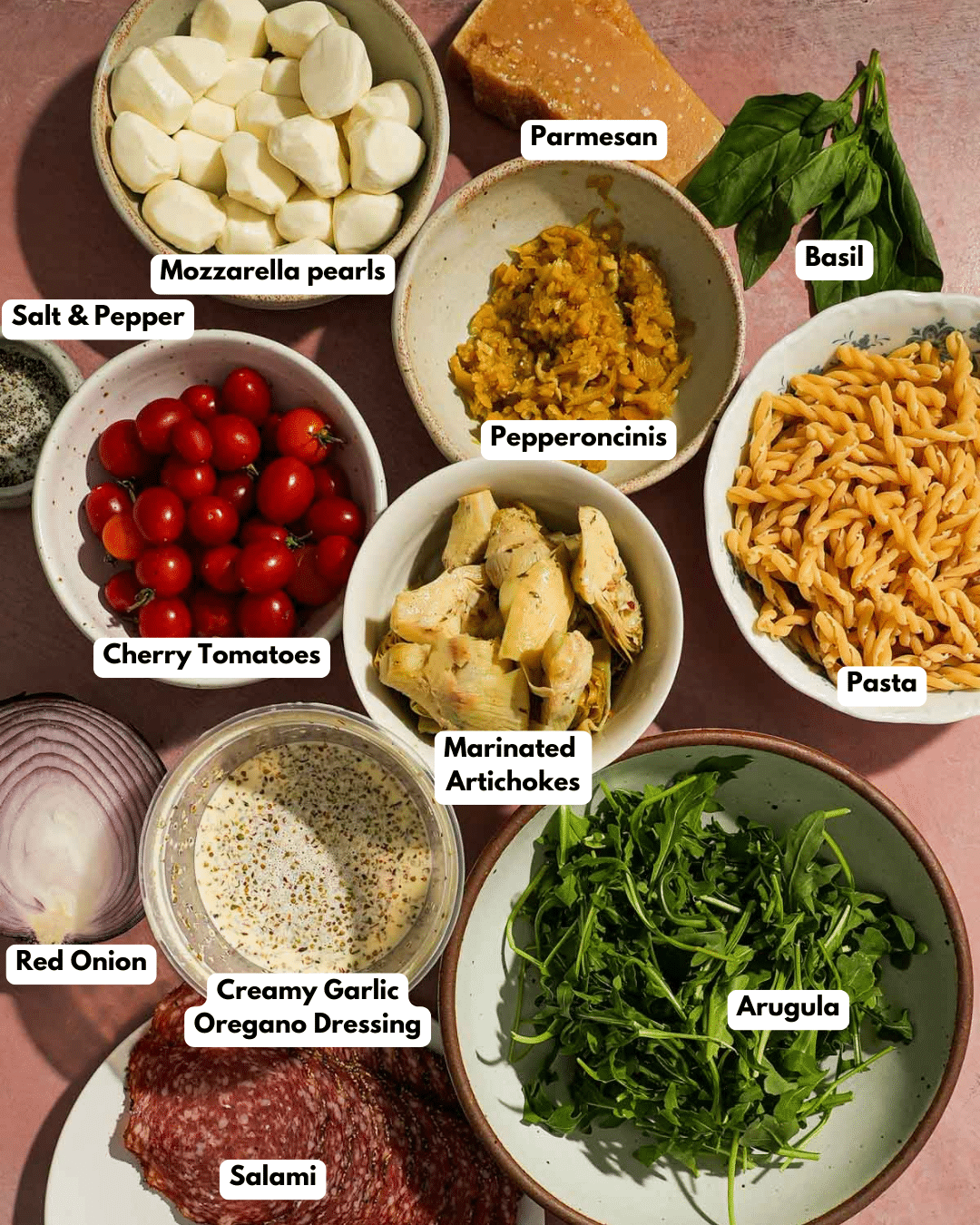 Pasta salad ingredients including mozzarella, tomatoes, arugula, salami, onions, and dressing.