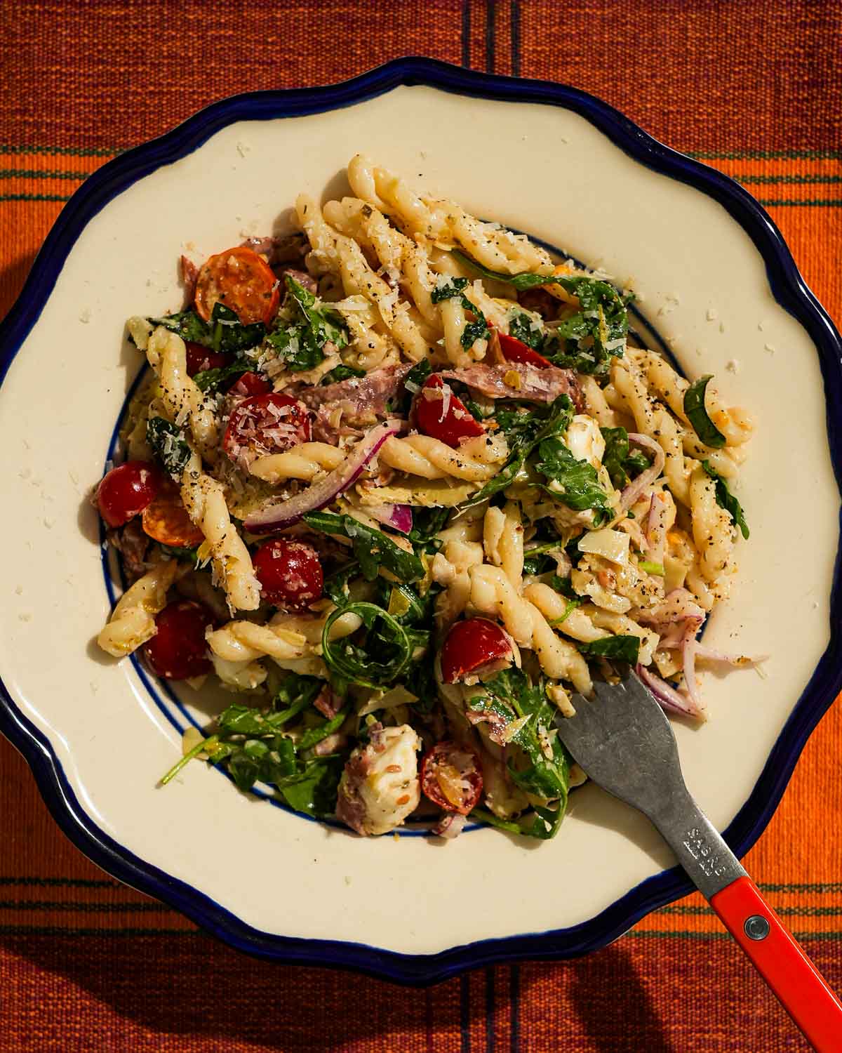 Pasta salad with tomatoes, arugula, salami, mozzarella, and dressing on plate.