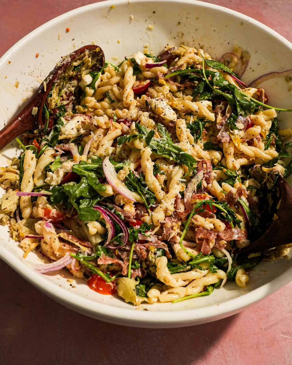 Large bowl of pasta salad mixed with greens, salami, tomatoes, and cheese.