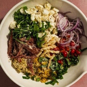 Bowl of pasta salad ingredients including arugula, mozzarella, onions, salami, and tomatoes.
