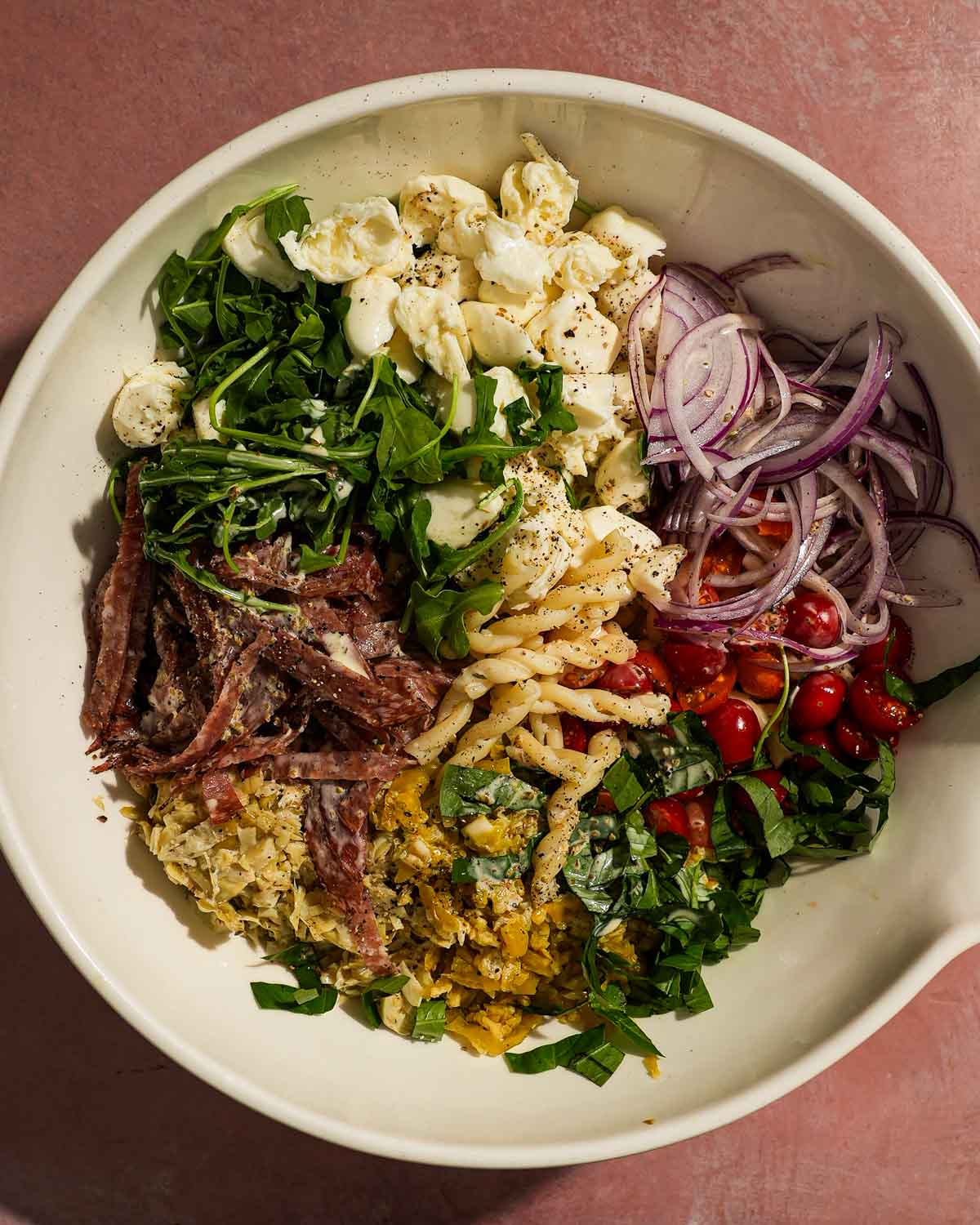 Unmixed pasta salad bowl with salami, mozzarella, onions, tomatoes, and chopped greens.