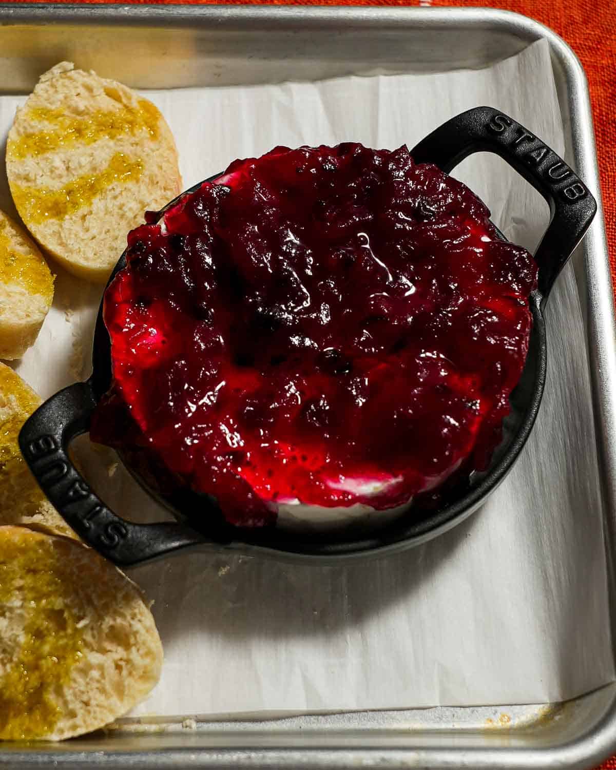 A sheet tray with parchment paper and baked brie with cranberry sauce and toast about to be put in the oven.