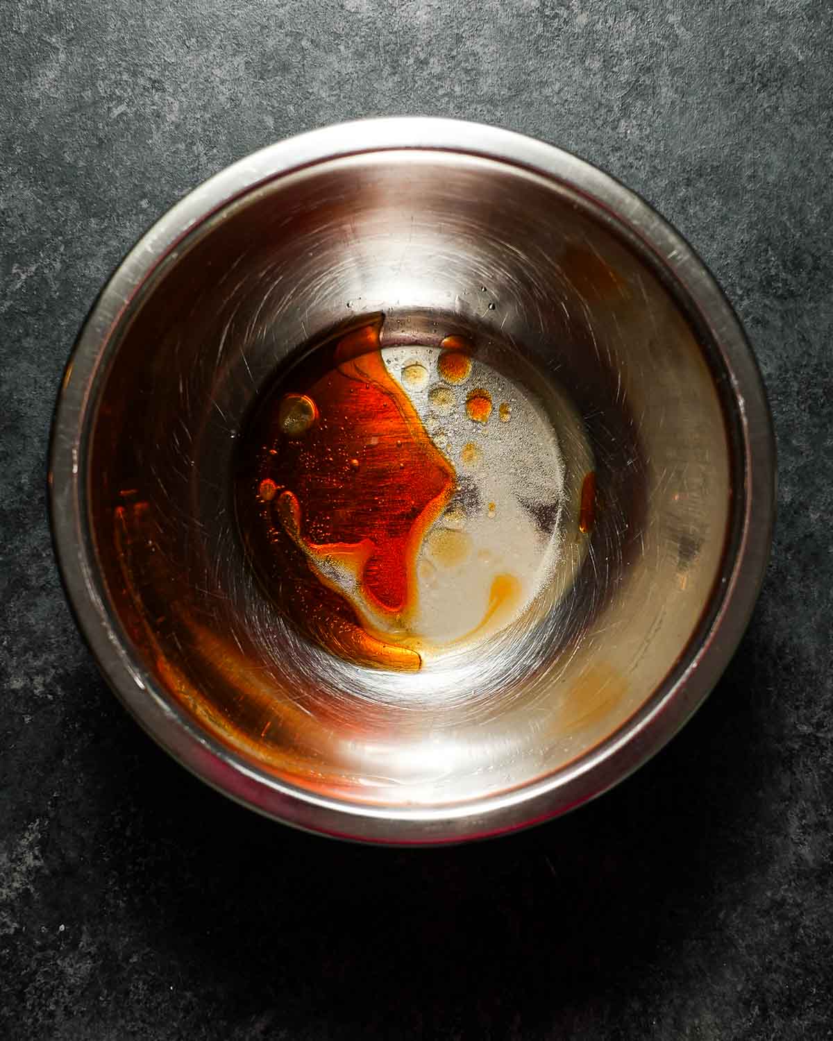 A metal bowl of a dressing of chili oil and vinegar, about to be tossed on cucumbers.