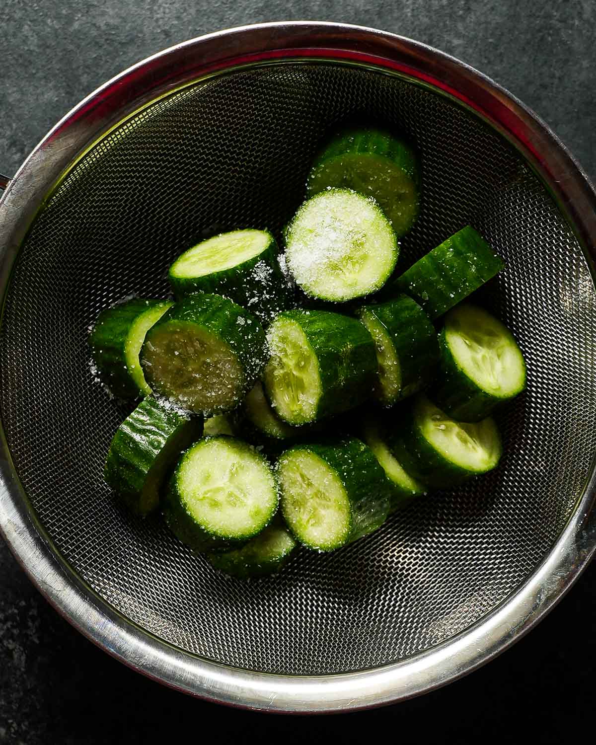 Sliced cucumber rounds over a colander with kosher salt and a bowl.