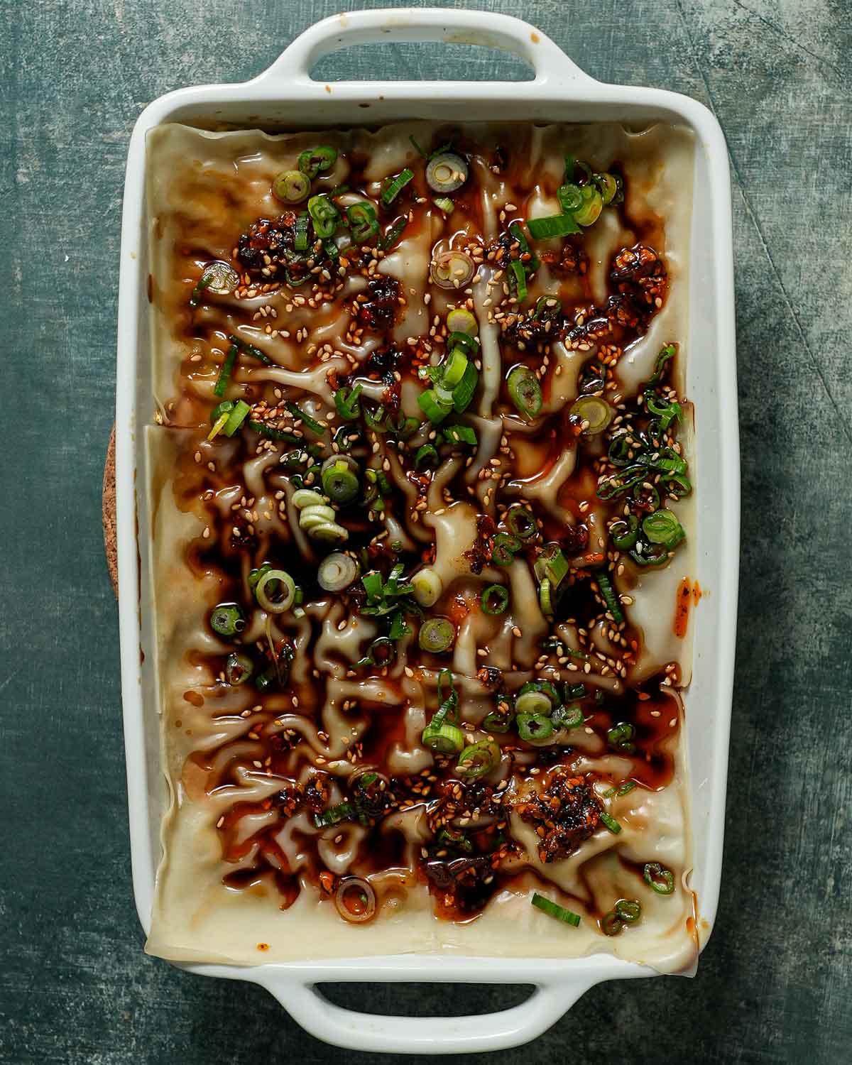 A baking pan layered with pork dumpling lasagna and soy sauce and chili oil as a topping.
