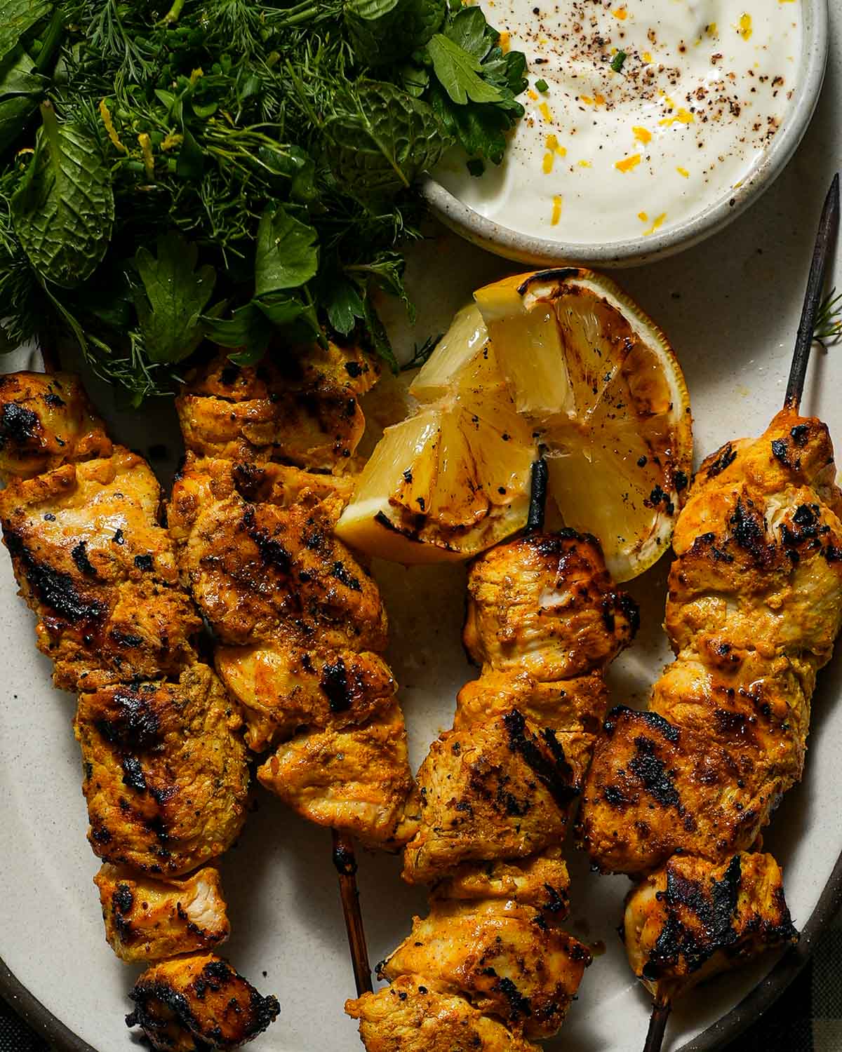 A plate of grilled yogurt marinated chicken with a charred lemon, a small bowl of yogurt and herbs.