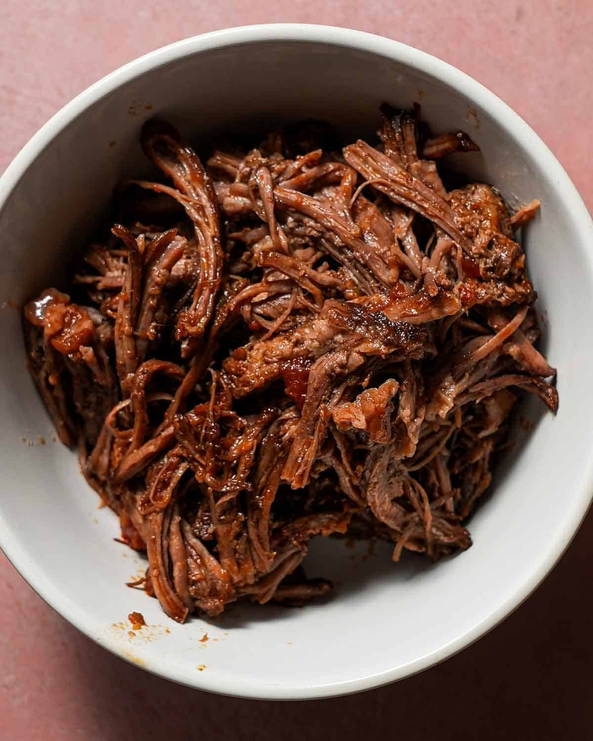 A white bowl of shredded short rib on a pink table.
