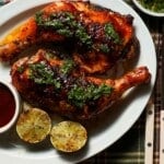 A platter of grilled honey chipotle bbq chicken with grilled limes and cilantro lime gremolata on a plaid tablecloth.