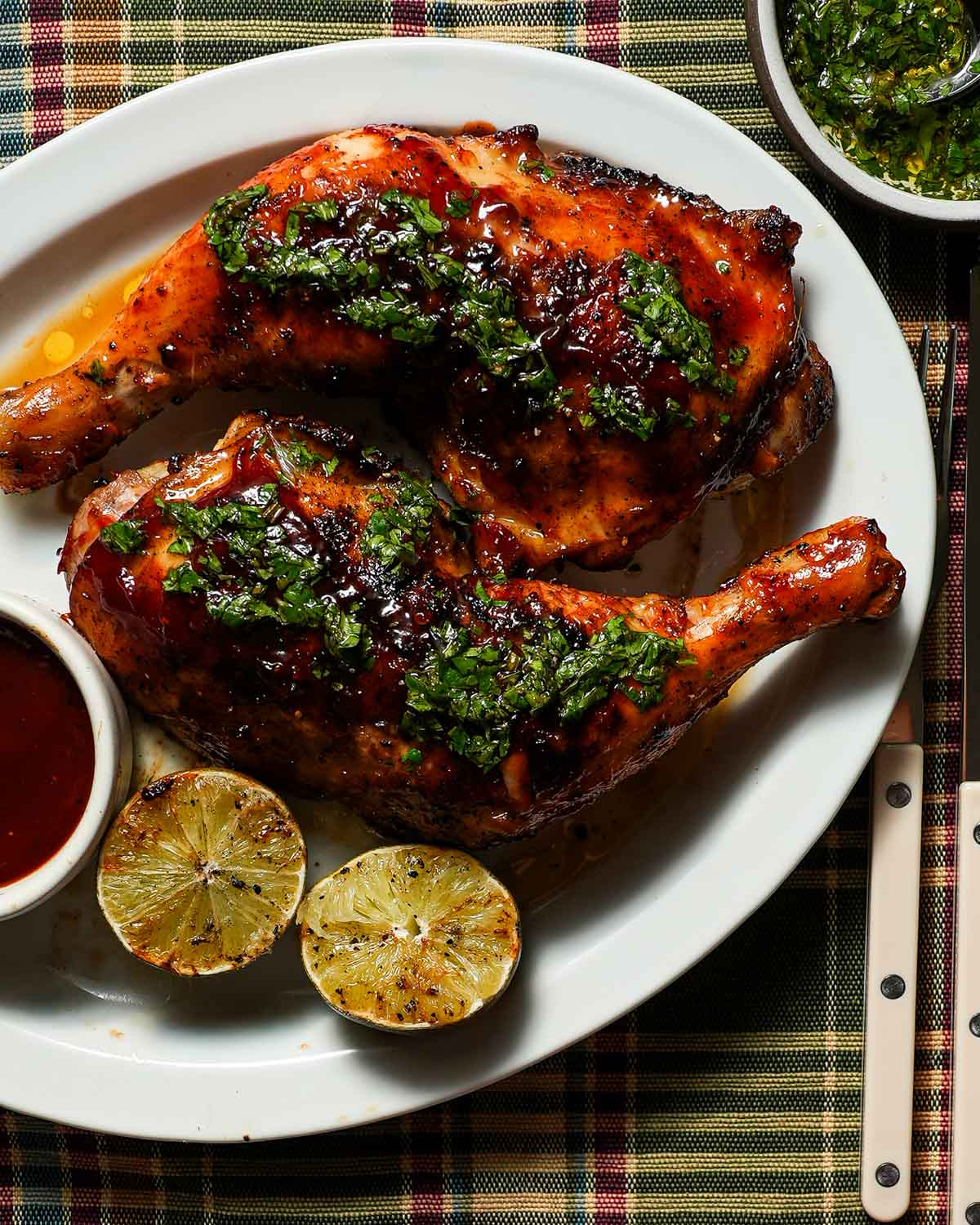 A platter of grilled honey chipotle bbq chicken with grilled limes and cilantro lime gremolata on a plaid tablecloth.