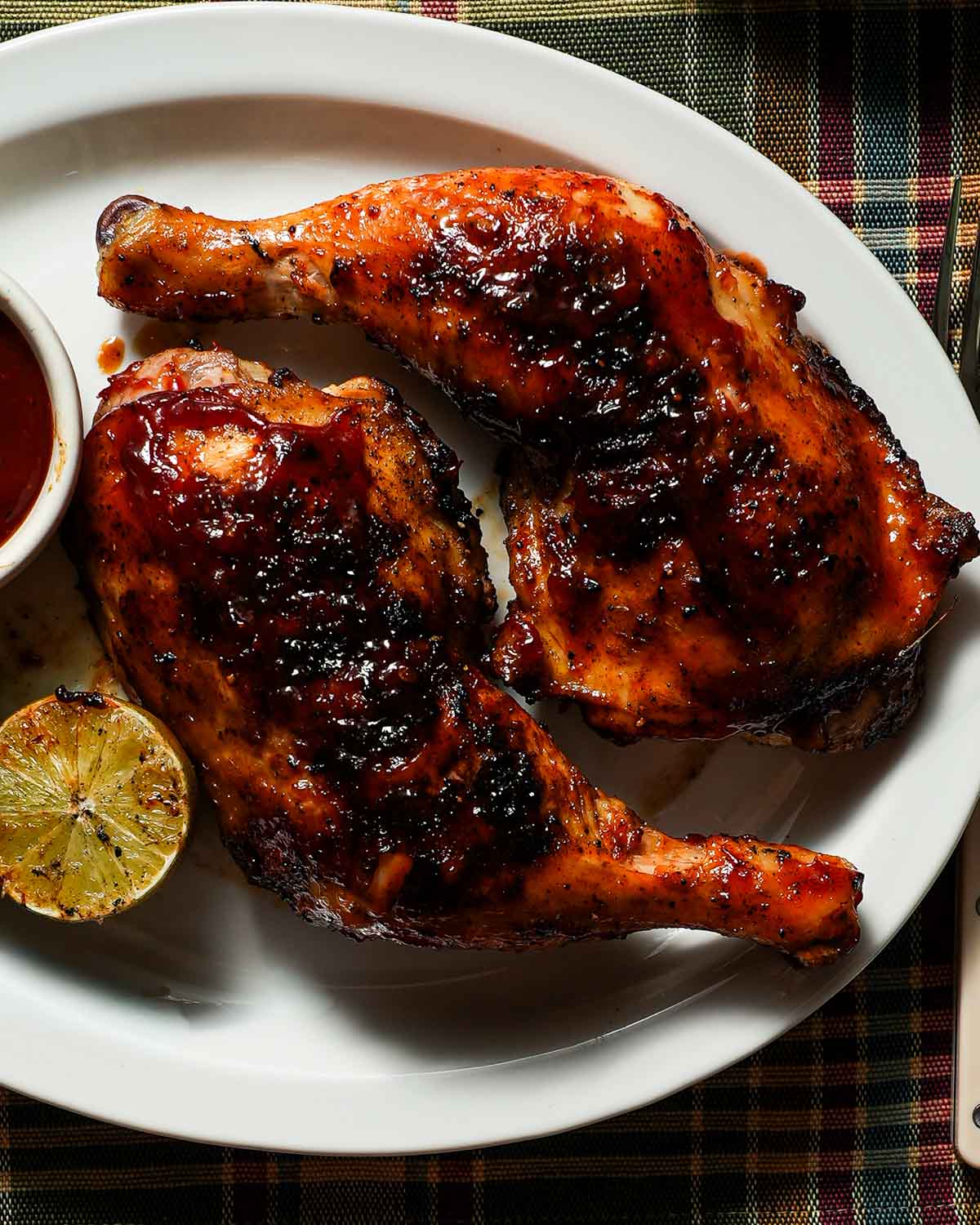 A white platter with 2 cooked chicken legs with bbq sauce, grilled lime and a side of barbecue sauce.