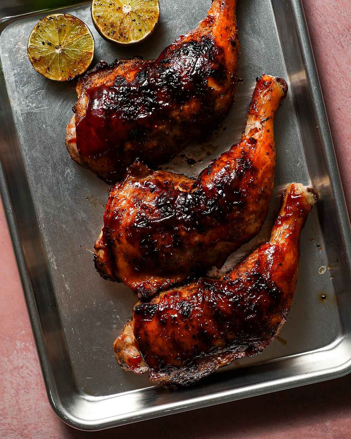 A baking sheet with 3 cooked chicken legs alongside halved grilled limes.