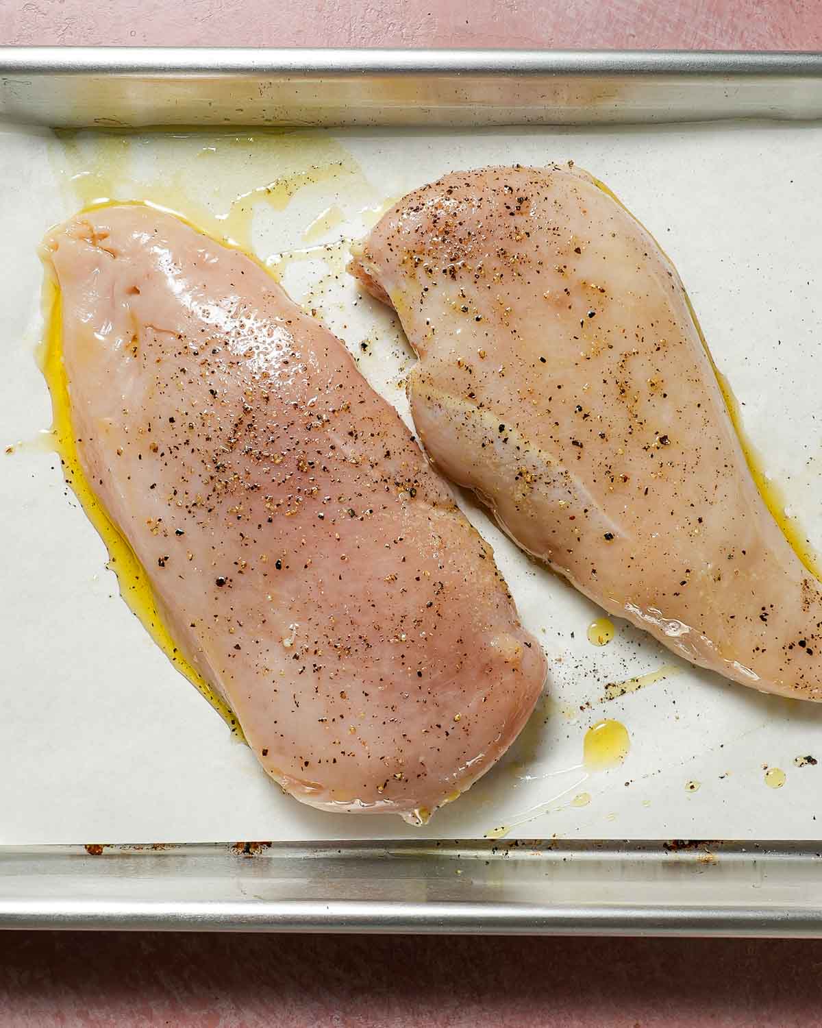 A baking sheet lined with parchment paper with raw chicken breasts with salt and pepper.