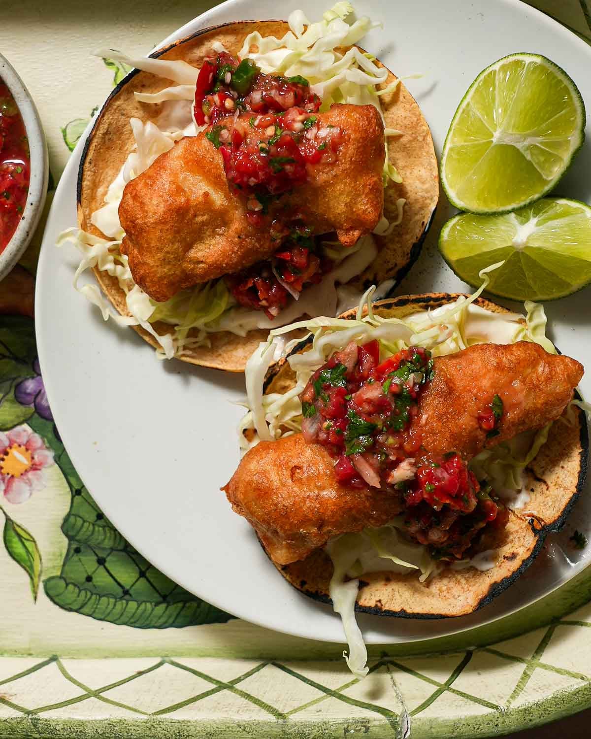 A plate of two crispy fish tacos with a lime wedge with tomato salsa.