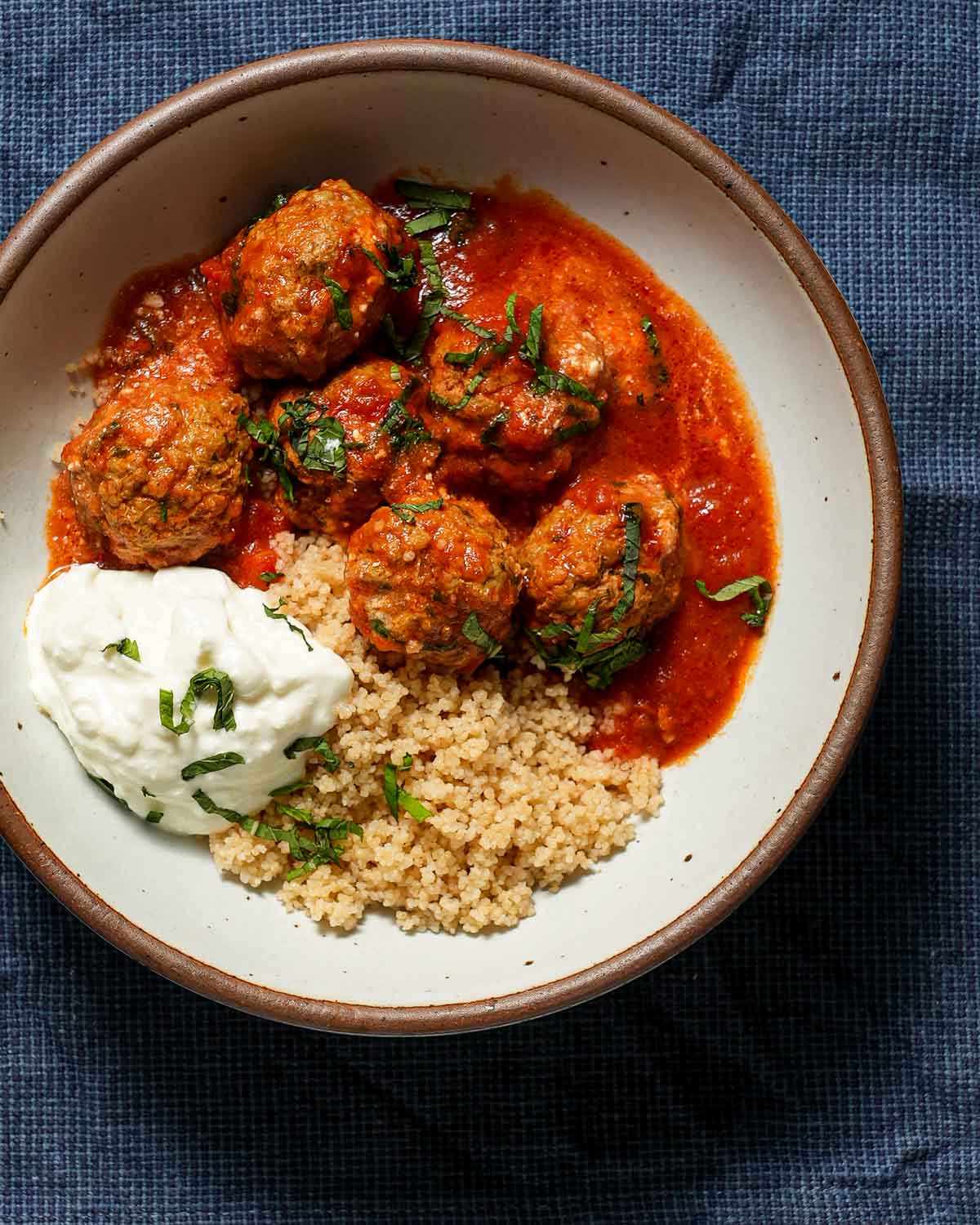 A bowl of lamb meatballs with yogurt, couscous and mint on a blue tablecloth.