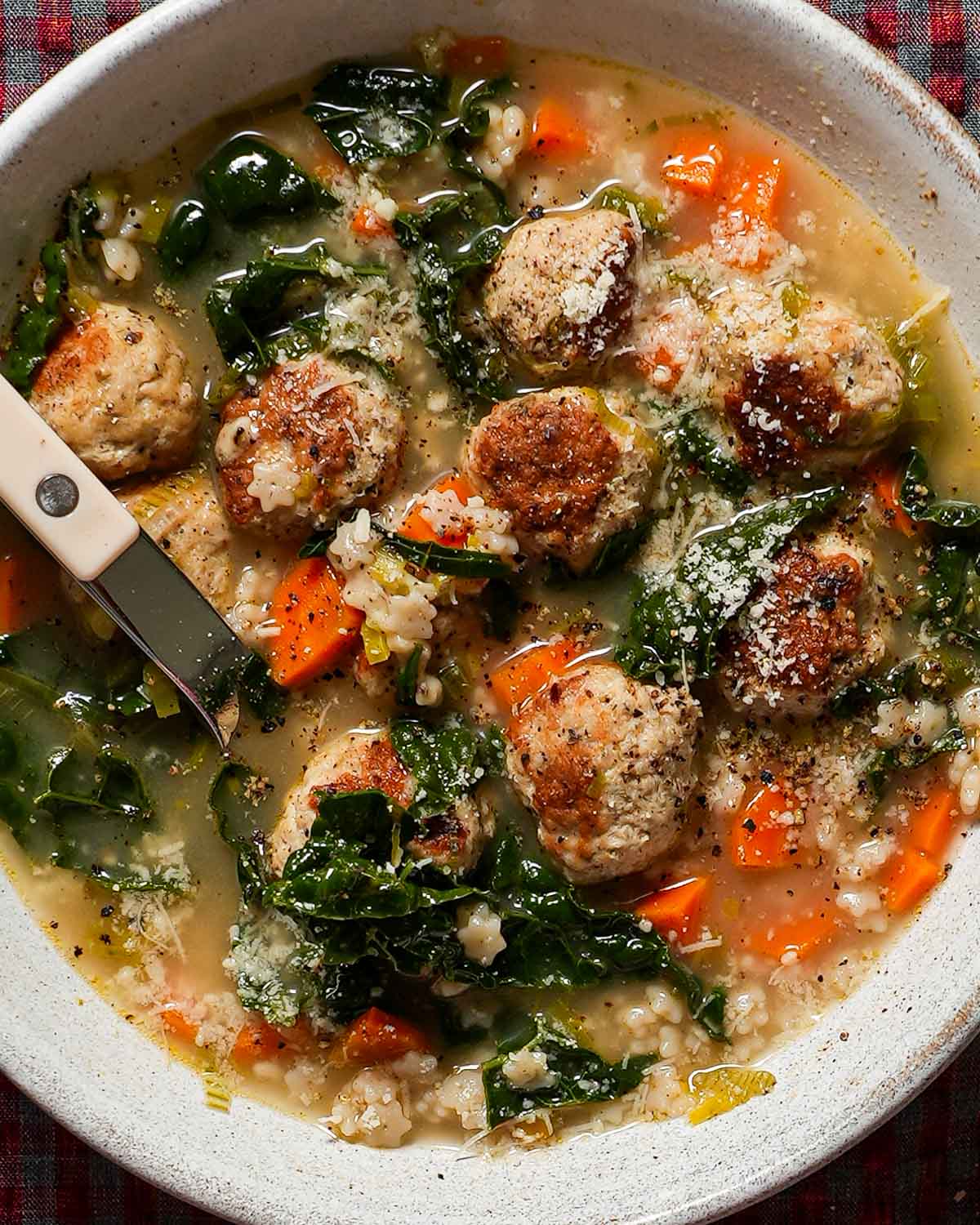 A bowl of meatball soup with a white bistro spoon, kale, and carrots.