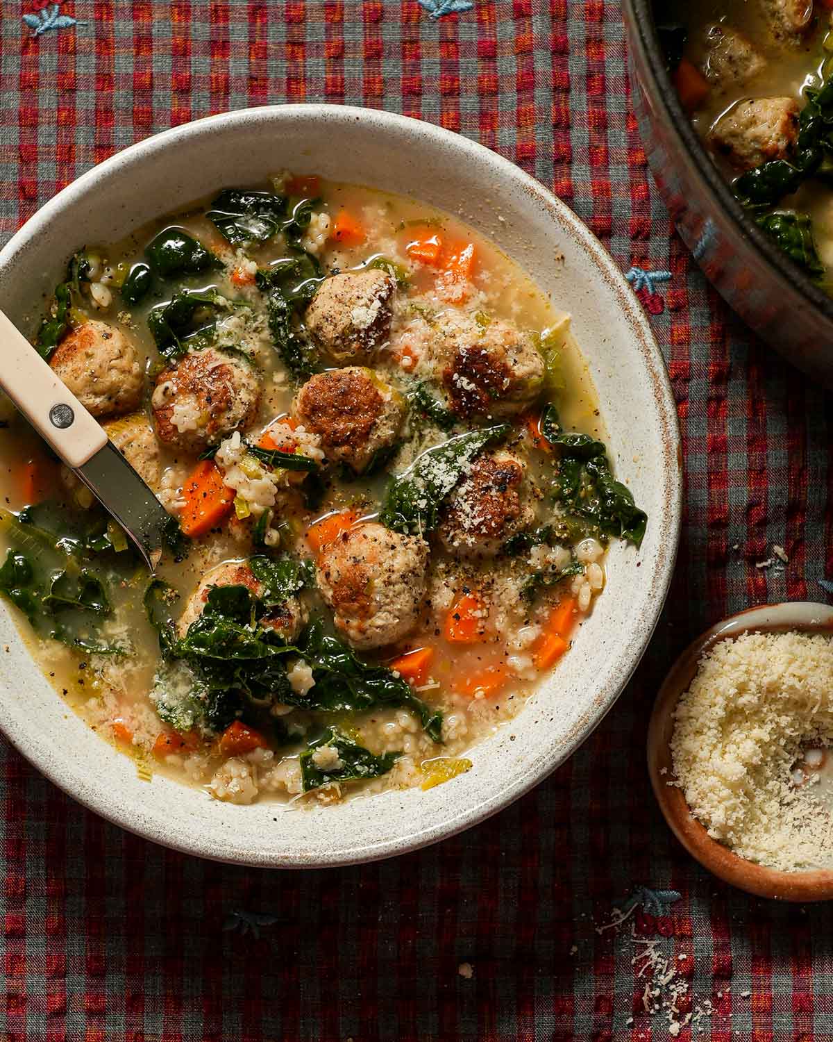 A bowl of Italian wedding soup with a bistro spoon and small bowl parmesan cheese next to it.