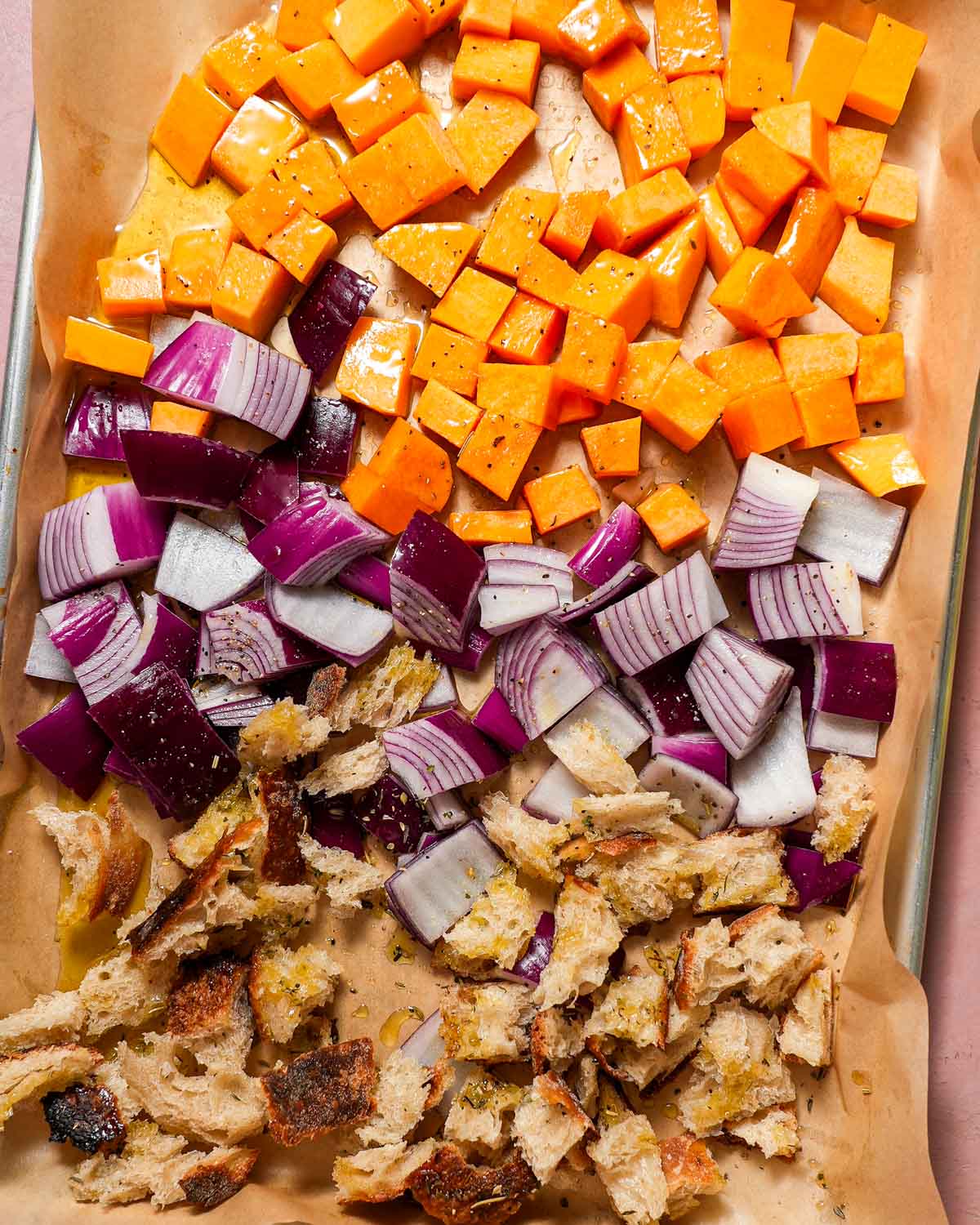 A baking sheet lined with parchment with butternut squash, red onions and croutons ready to be roasted.